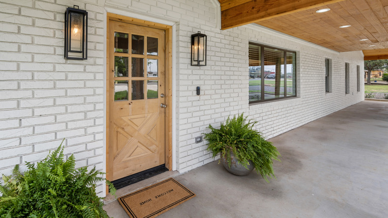 A white brick home with a wood front door, lighting sconces, and ferns