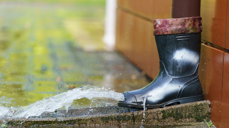 A black rain boot at the bottom of a downspout, with its toes cut out so water can flow through