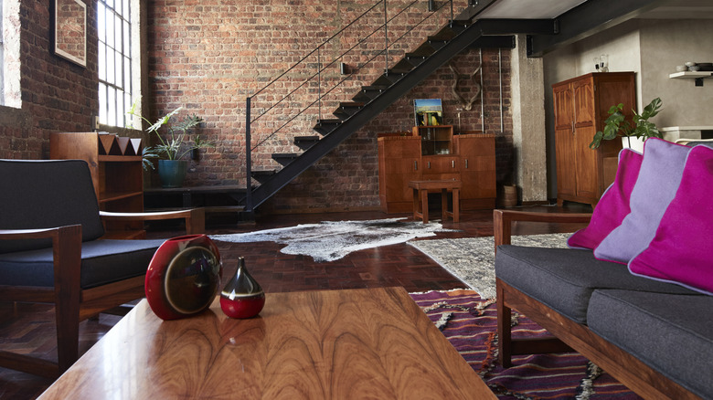 A modern living room with a brick wall and metal staircase with blue cushioned wood furniture.