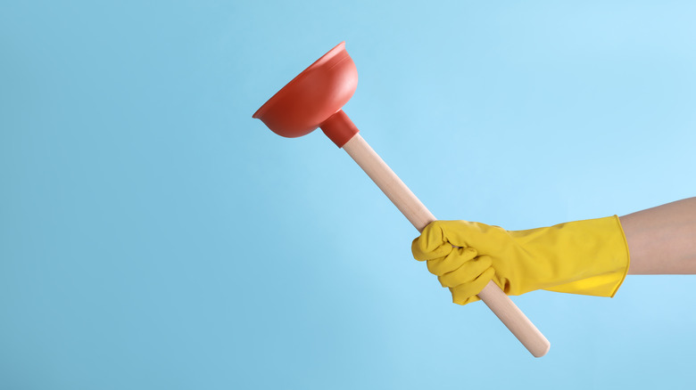woman wearing yellow gloves holding a toilet plunger against a blue background
