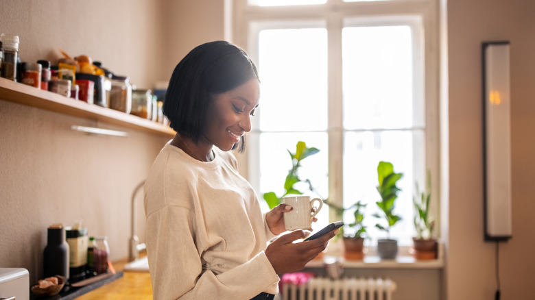 Woman stands in kitchen with coffee cup in one hand checking her phone