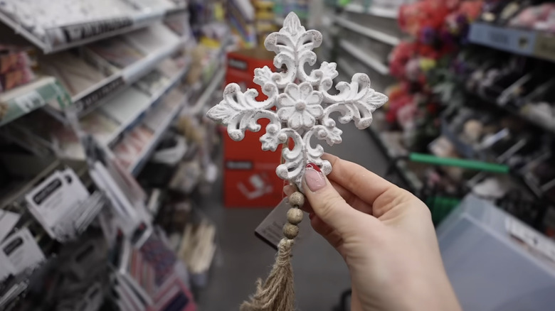A person holds a small, ornate white wooden decoration with a natural wood bead tassel in a Dollar Tree store aisle
