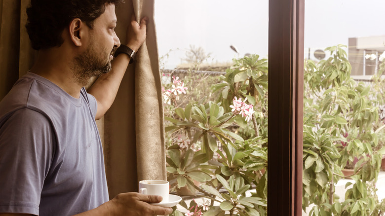 A man holding a cup of coffee holds back a curtain to look out a window.