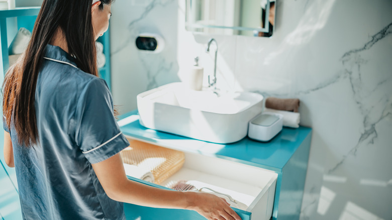 A hotel guest looks into an open vanity drawer in the bathroom while wearing blue silk pajamas.