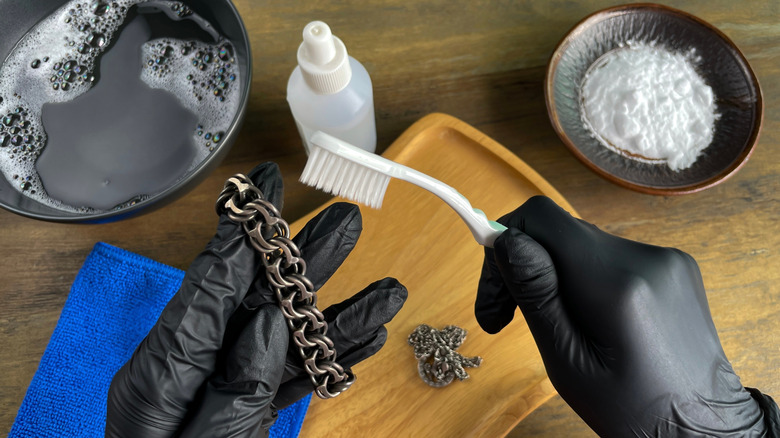 Gloved hands holding a tarnished silver chain to clean with lots of supplies including toothbrush