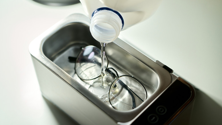 An unseen hand pours distilled water into an ultrasonic cleaner to wash eyeglasses