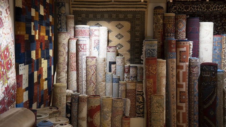 Many rugs rolled up and hung on the wall of a shop, showing a wide variety of colors, sizes, and patterns