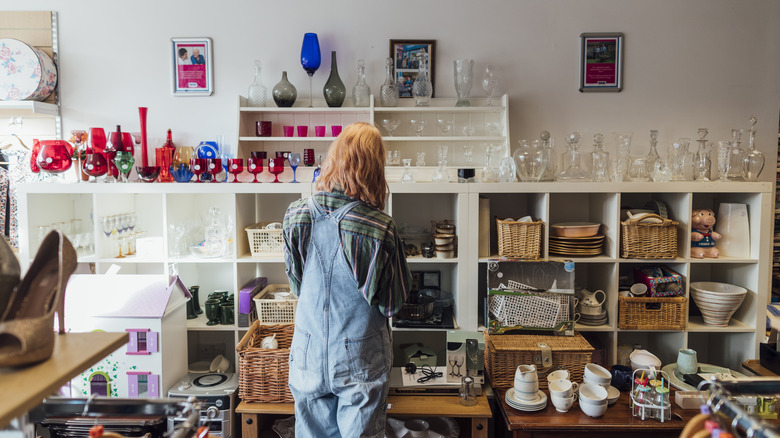 A person with shoulder-length dark blond hair wearing light blue jean overalls and a striped shirt is looking at a wall of white shelves that are full of kitchenware and household items.