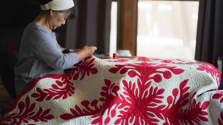 A woman wearing a white handband and a long-sleeved shirt is stitching a red and white quilt with a botanical motif.