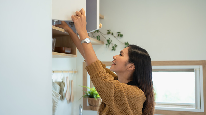 A woman reaches up to a high cabinet to retrieve something