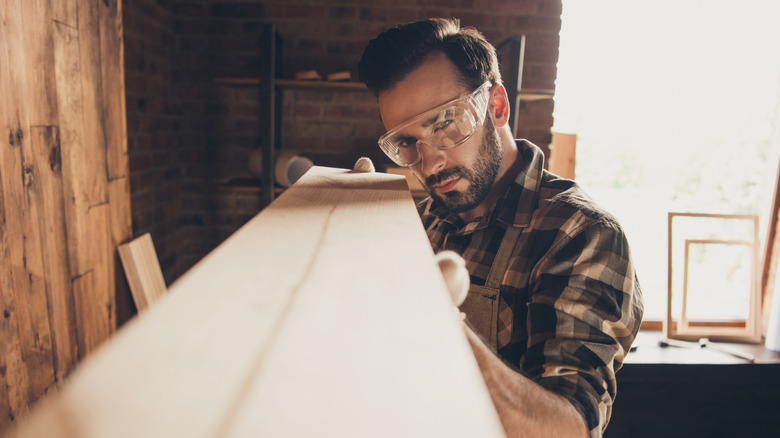 A man with safety glass and a brown checked shirt checking a wooden shelf for imperfections.