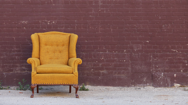 A golden vintage wingback chair sitting outside in front of a red brick wall