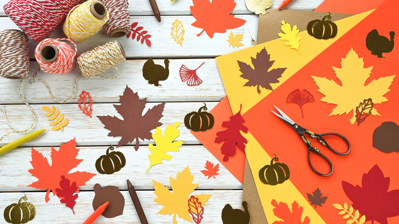 Craft supplies on a table, including paper cutouts of leaves, pumpkins, and turkey, as well as spools of colored twine and a pair of scissors
