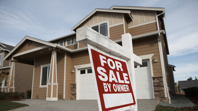 A red 'For Sale By Owner' sign sits in front of a brown house
