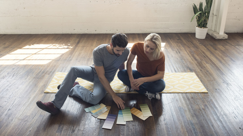 Two people inspecting paint swatches while sitting on dark-colored wood floors.