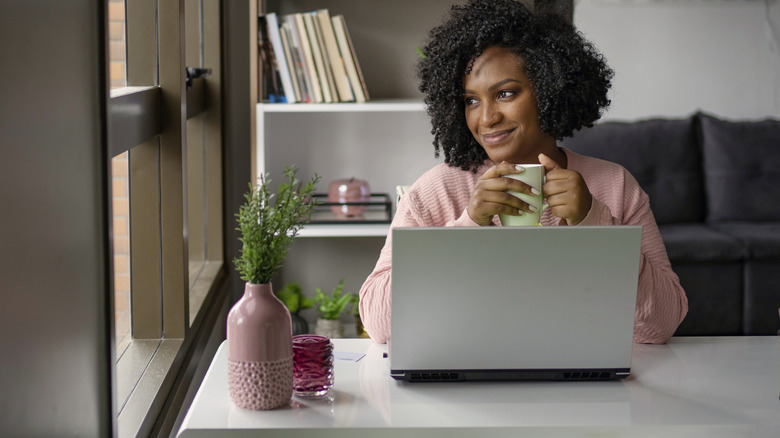 Woman drinking out of a mug while using laptop at home