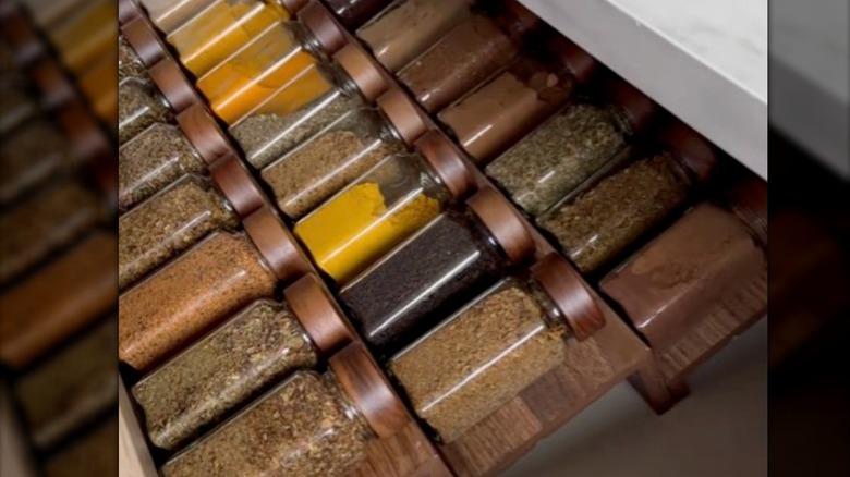 Glass spice jars with wood-colored lids sitting in a drawer on a riser made from an IKEA picture ledge