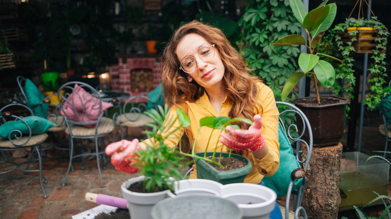 A woman with glasses and a yellow shirt sits outdoors with houseplants and potting materials in front of her.