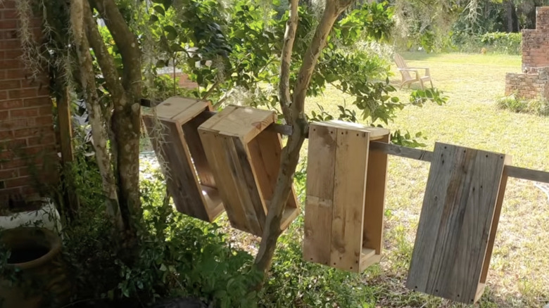 Nuc boxes made from wood pallets hang from a wooden beam in a shaded area of a residential backyard.
