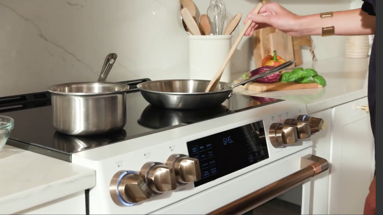 Person cooking on the Cafe double induction range with silver pots and a woman wearing a thick bracelet and stirring with a wooden spoon.