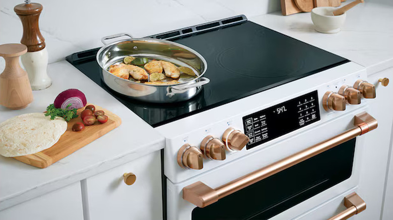 White Cafe 30-inch double induction range with brushed bronze knobs, a silver pan on top, and food to the side