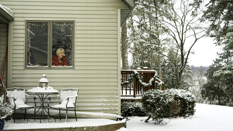 A woman looks out the window of a home onto a backyard covered in snow.