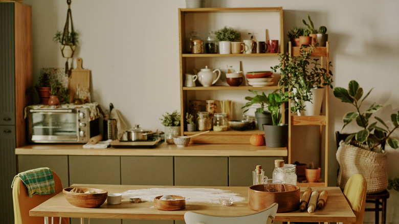 A kitchen with a packed counter and shelves, dishes, appliances, and plants as well as a table with flour, pans, and rolling pins.