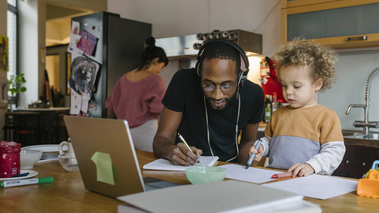 A man wearing headphones works at a desk in a kitchen while his son plays beside him and his wife cooks behind him