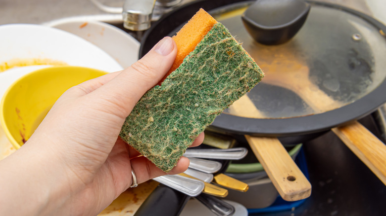 A person holds a dirty old kitchen sponge over some dirty dishes.