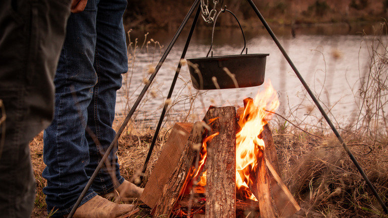 A cast iron pot hanging over an open fire next to legs wearing cowboy boots