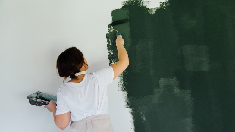 A woman painting a white wall deep green, holding a paint tray and a paint roller