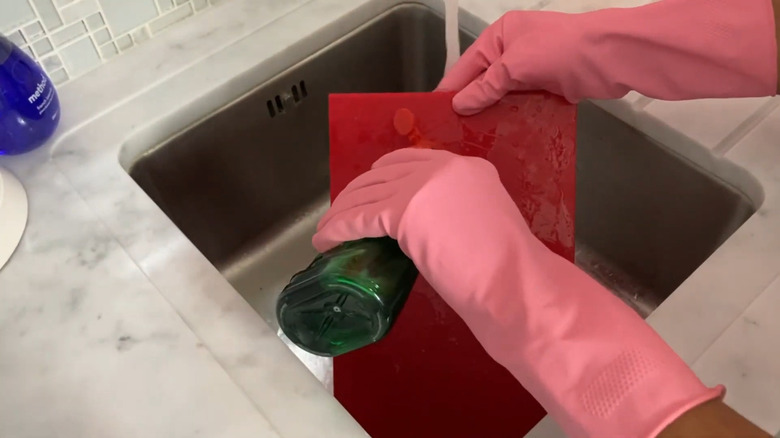 Person with pink rubber gloves washing a red plastic cutting board with dish soap in a sink