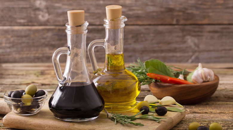 Bottles of vinegar and oil sitting on a wooden table next to olives, garlic cloves, and herbs