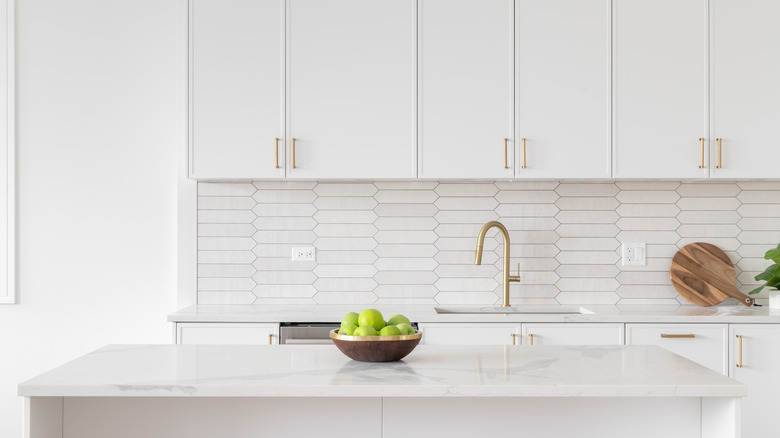 A beautiful kitchen faucet detail with white cabinets, a gold faucet, white marble countertops, and a brown picket ceramic tile backsplash.