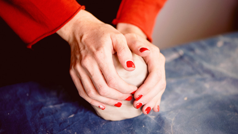 A pair of light-skinned hands sporting bright red nails knead a ball of clay.
