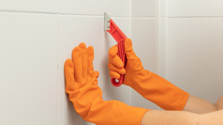 Person's hands with orange gloves holding a grout removal tool against white tile walls.