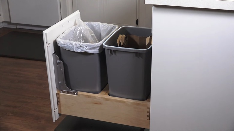 A kitchen cabinet with a trash can drawer open revealing two cans with bright yellow bags
