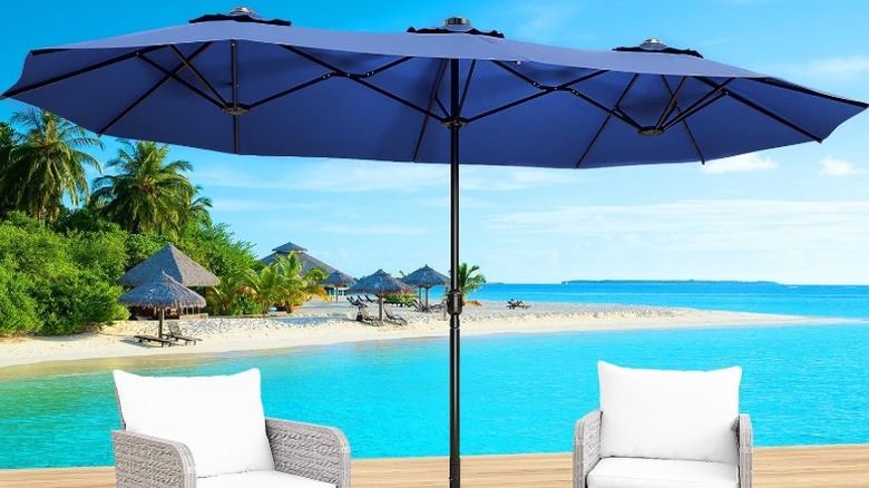 The large patio umbrella on a wooden deck next to the beach with two white outdoor armchairs underneath it