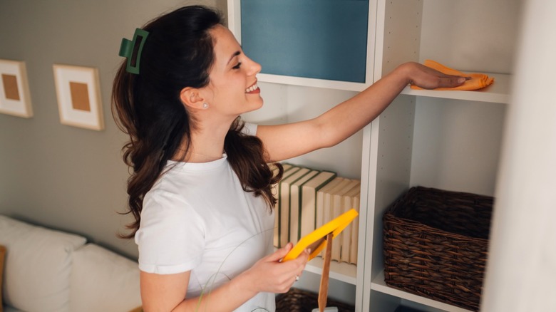 smiling woman dusting shelves with orange cloth while holding picture frame