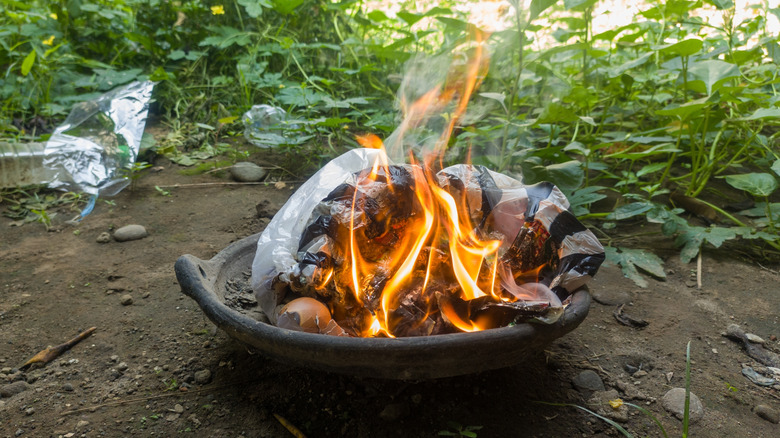 A trash fire burning in cast iron bowl on a patch of bare dirt in a backyard.