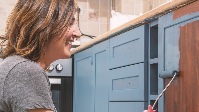 A smiling woman painting her kitchen cabinets light blue with a roller