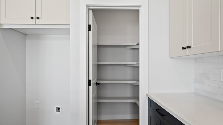 An open door in a kitchen reveals an empty white walk-in pantry with no lights