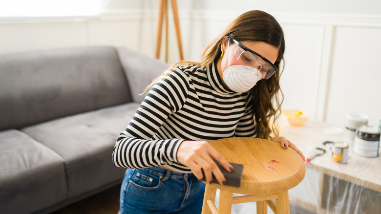 A young woman wears a face mask and goggles to work on a wooden stool.