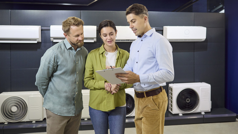 A salesman talking with a man and a woman with air-conditioners behind them.