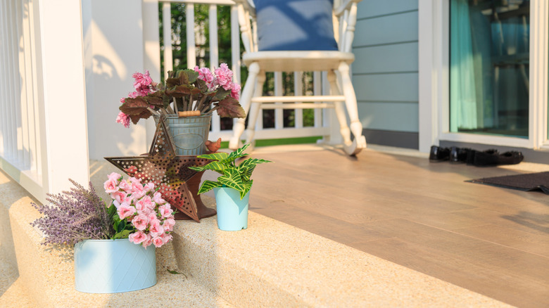 Artificial flowers and foliage in planters on porch stairs with a metal outdoor sculpture and a rocking chair in the background.