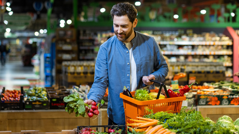 Man in blue shirt shopping in a grocery store, he holds a bunch of radishes in one hand, full basket in the other
