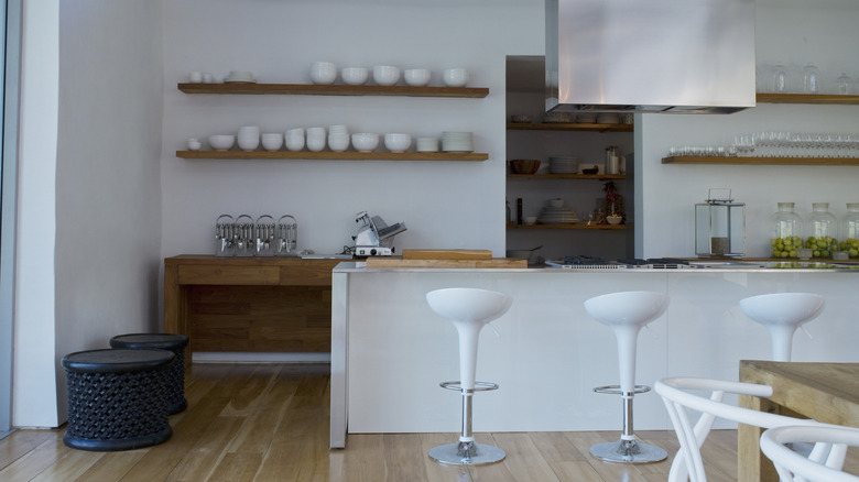 White kitchen with three white bar stools in the foreground, open wood shelving stacked with white bowls in the background, and wood counters with kitchen tools on them.