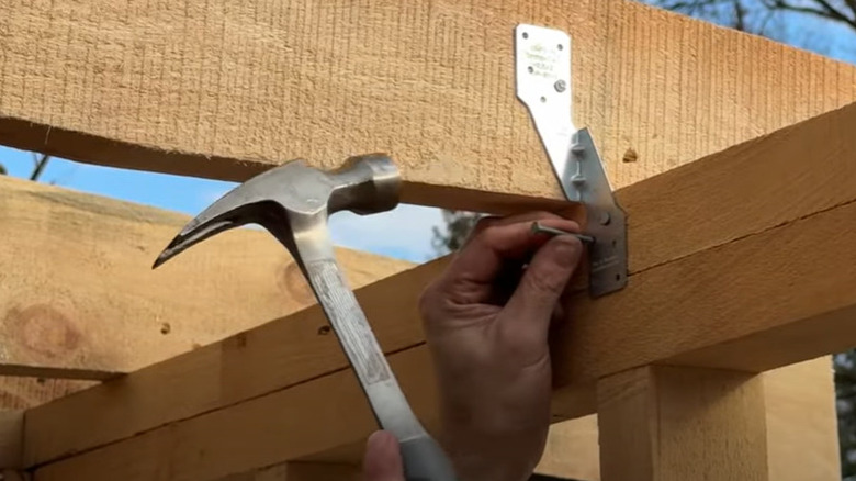 A person holding a nail and hammer while fastening a metal hurricane strap to wooden roof framing