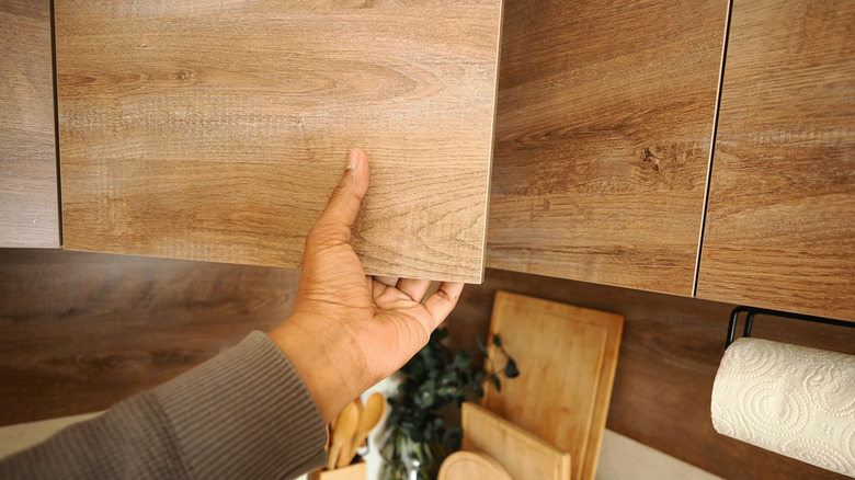A person's hand opening a wooden cabinet door in a kitchen space.