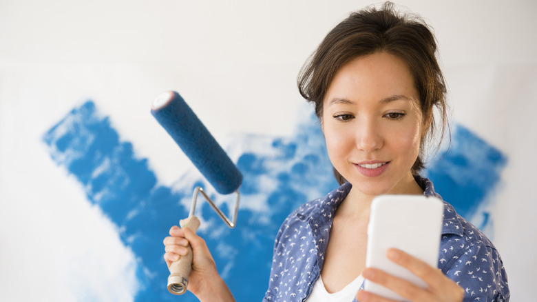 A girl looks at her phone as she prepares to paint a wall with blue paint
